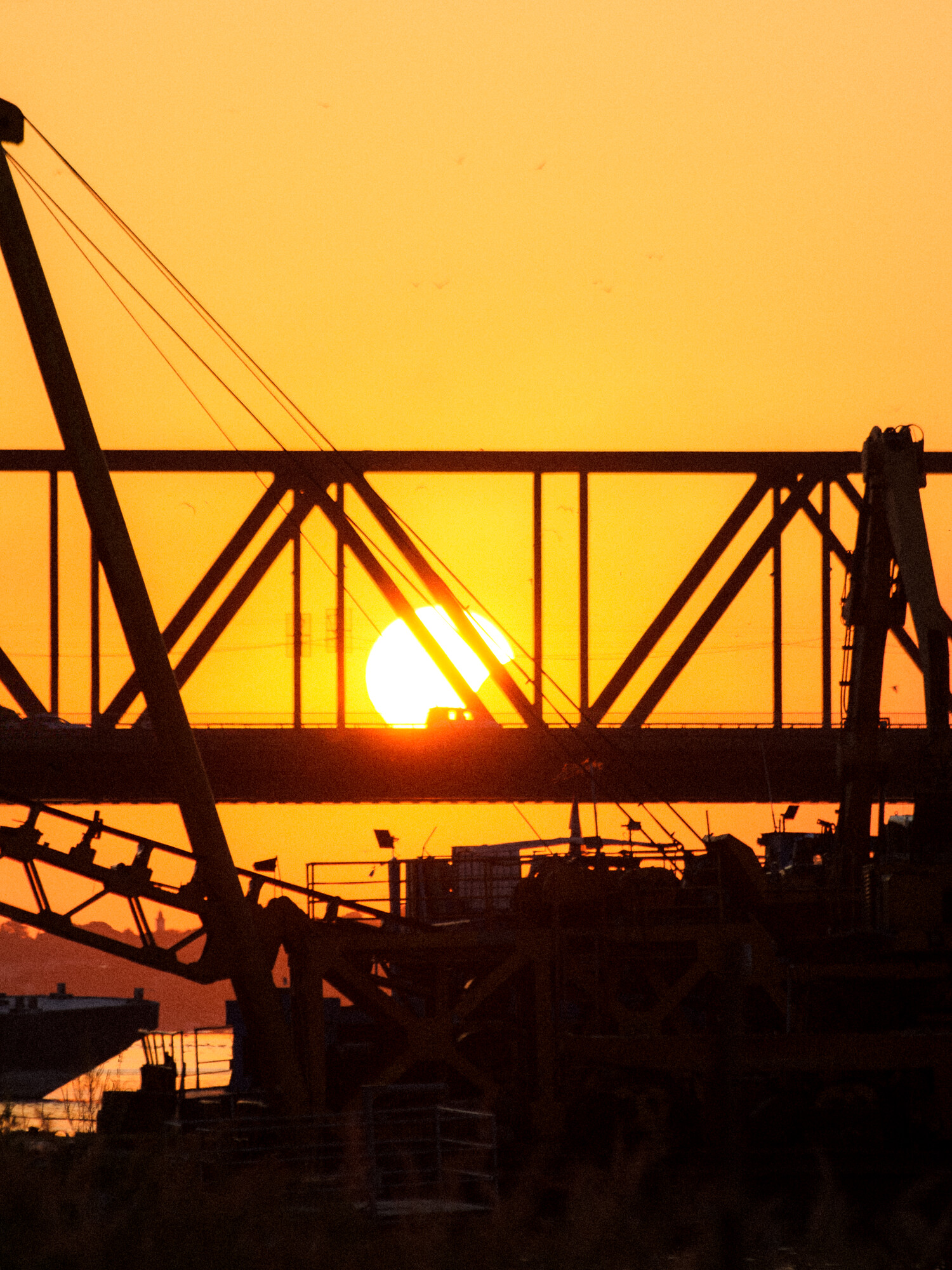 Partially eclipsed sun setting behind a truss bridge, silhouetted against orange sky with cranes in foreground