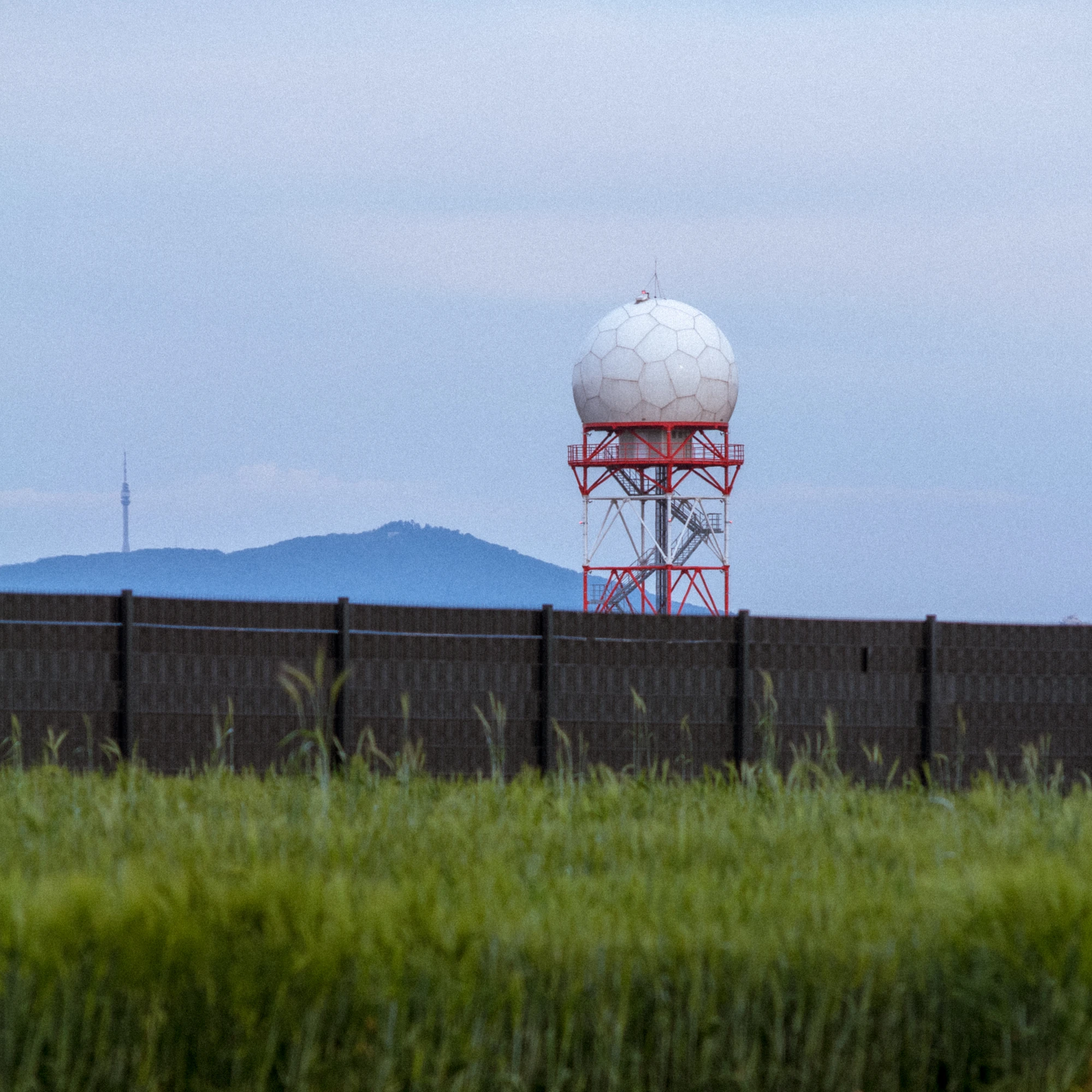 Radar tower behind a fence with grassy field and cloudy hills in the background