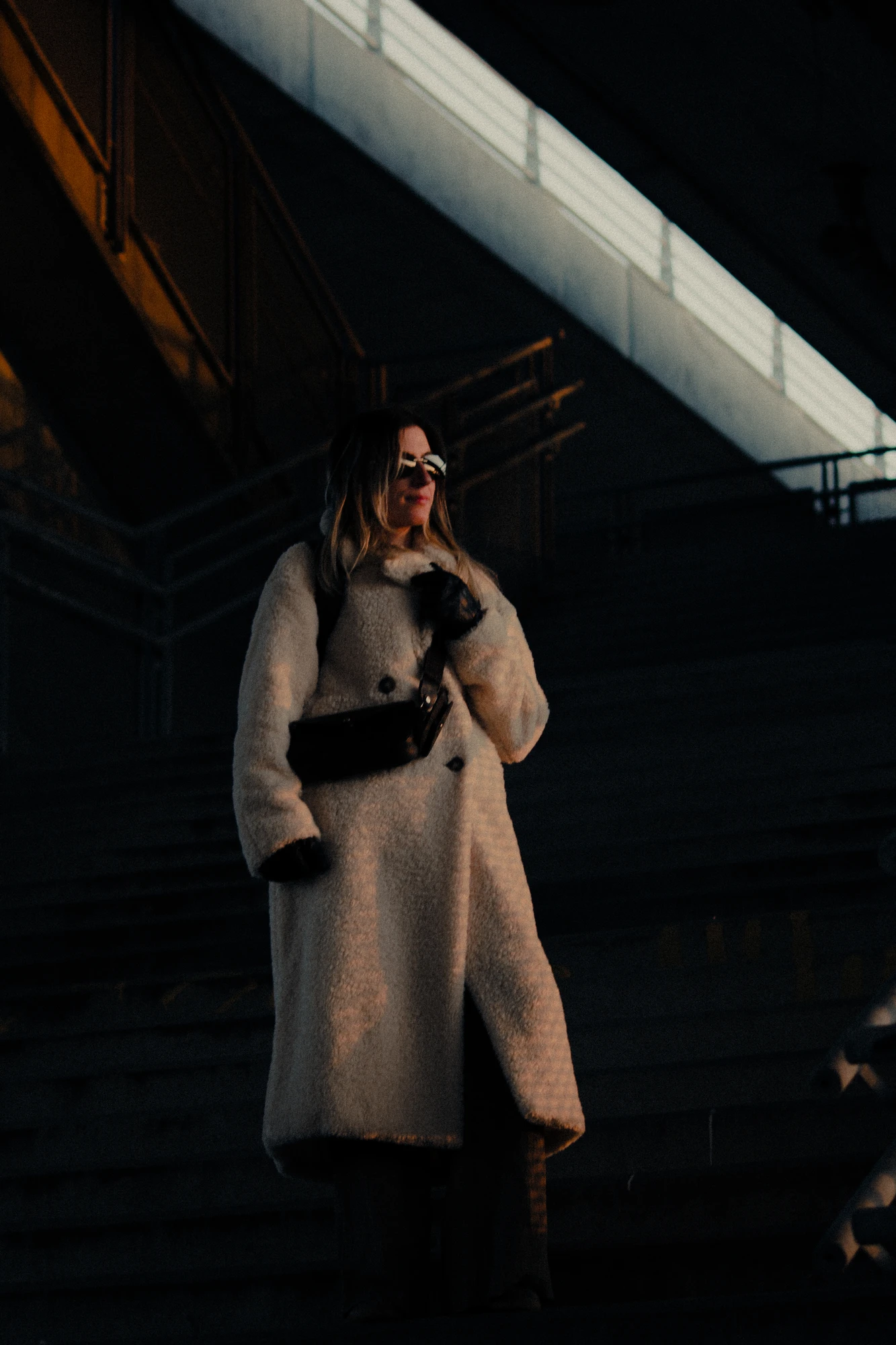 Woman in white coat and sunglasses standing on the stairs under a bridge