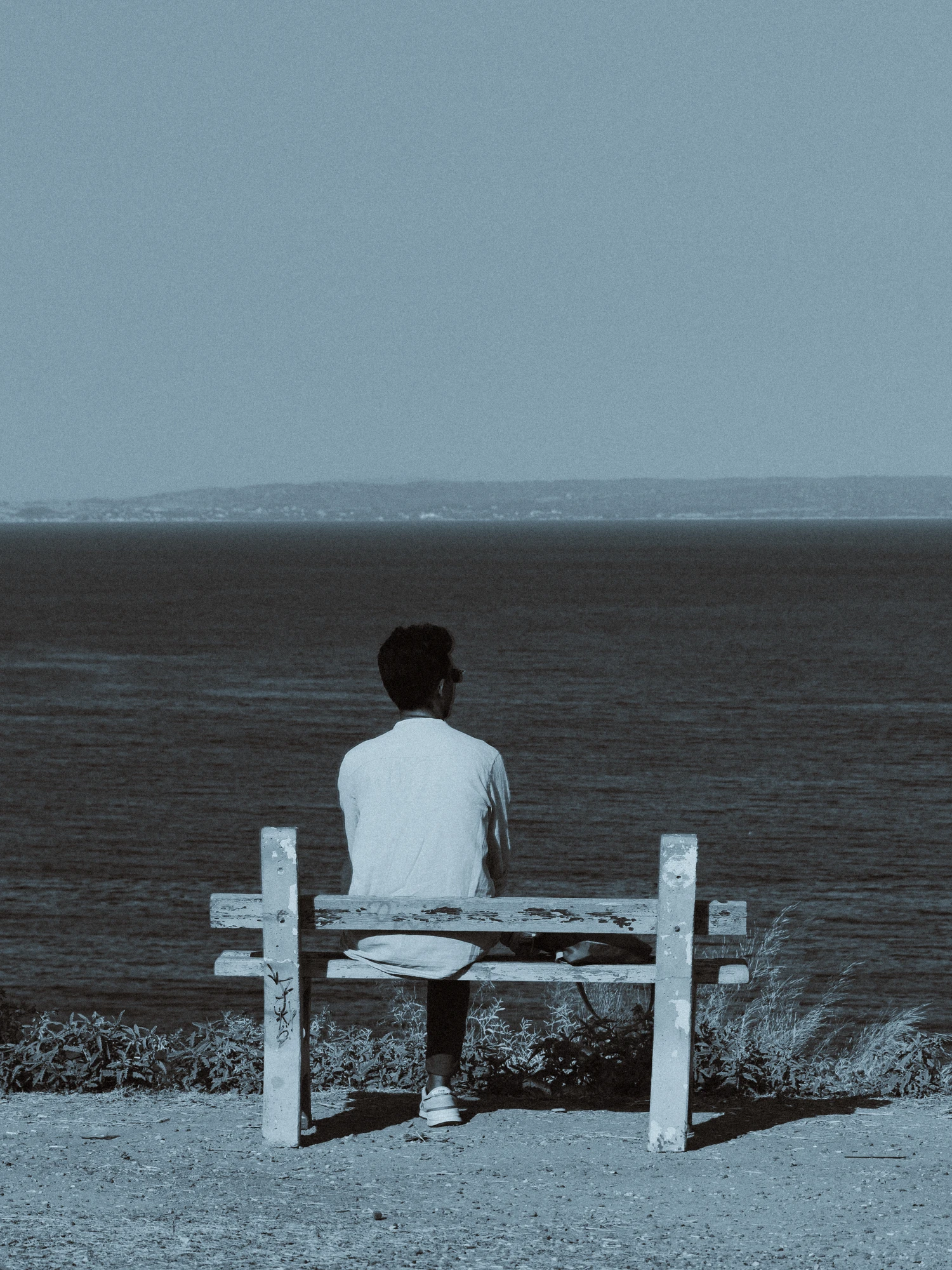 Person sitting alone on a weathered bench overlooking the sea toward distant hills
