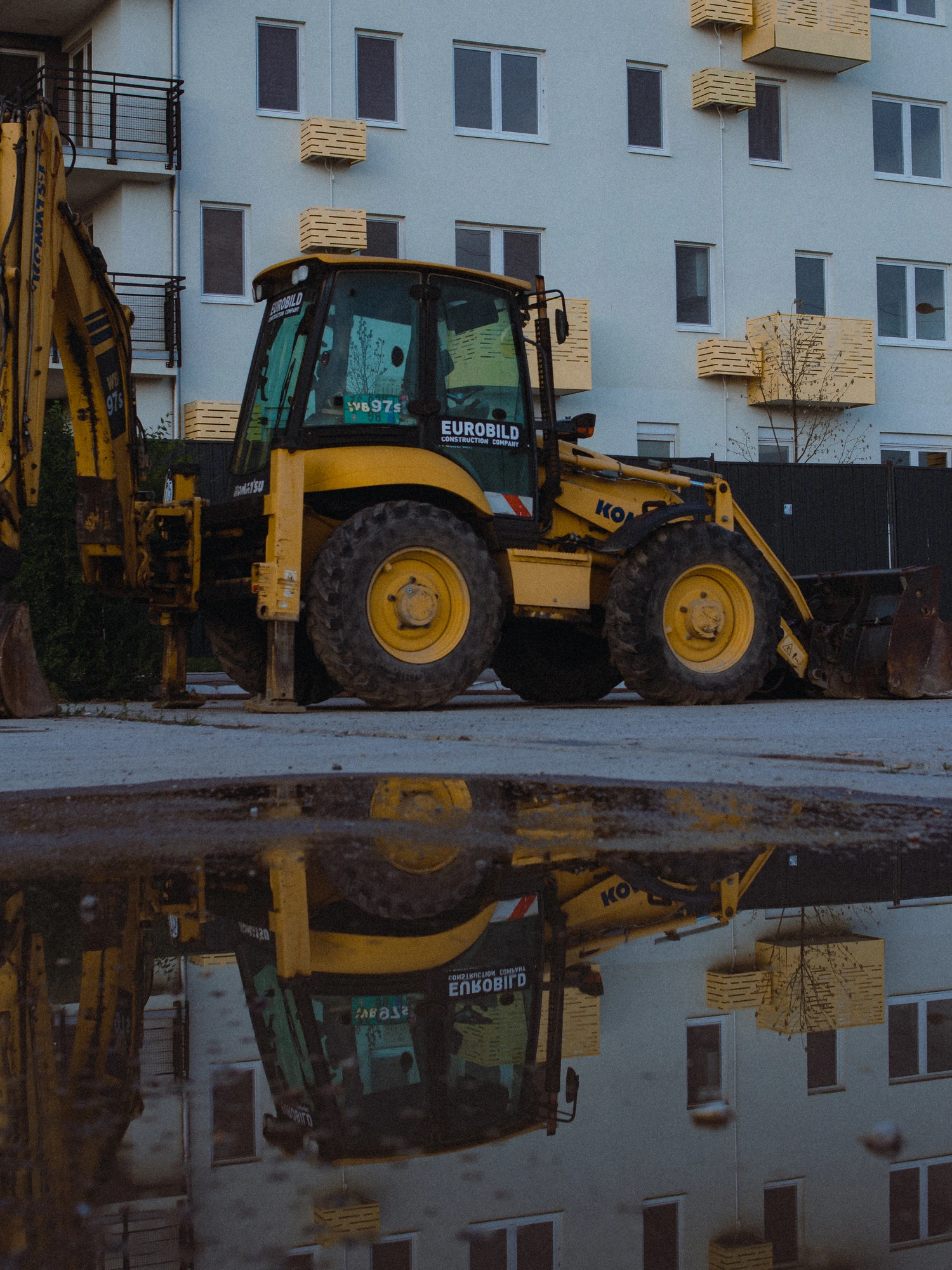Yellow backhoe loader parked beside an apartment building, reflected in a puddle