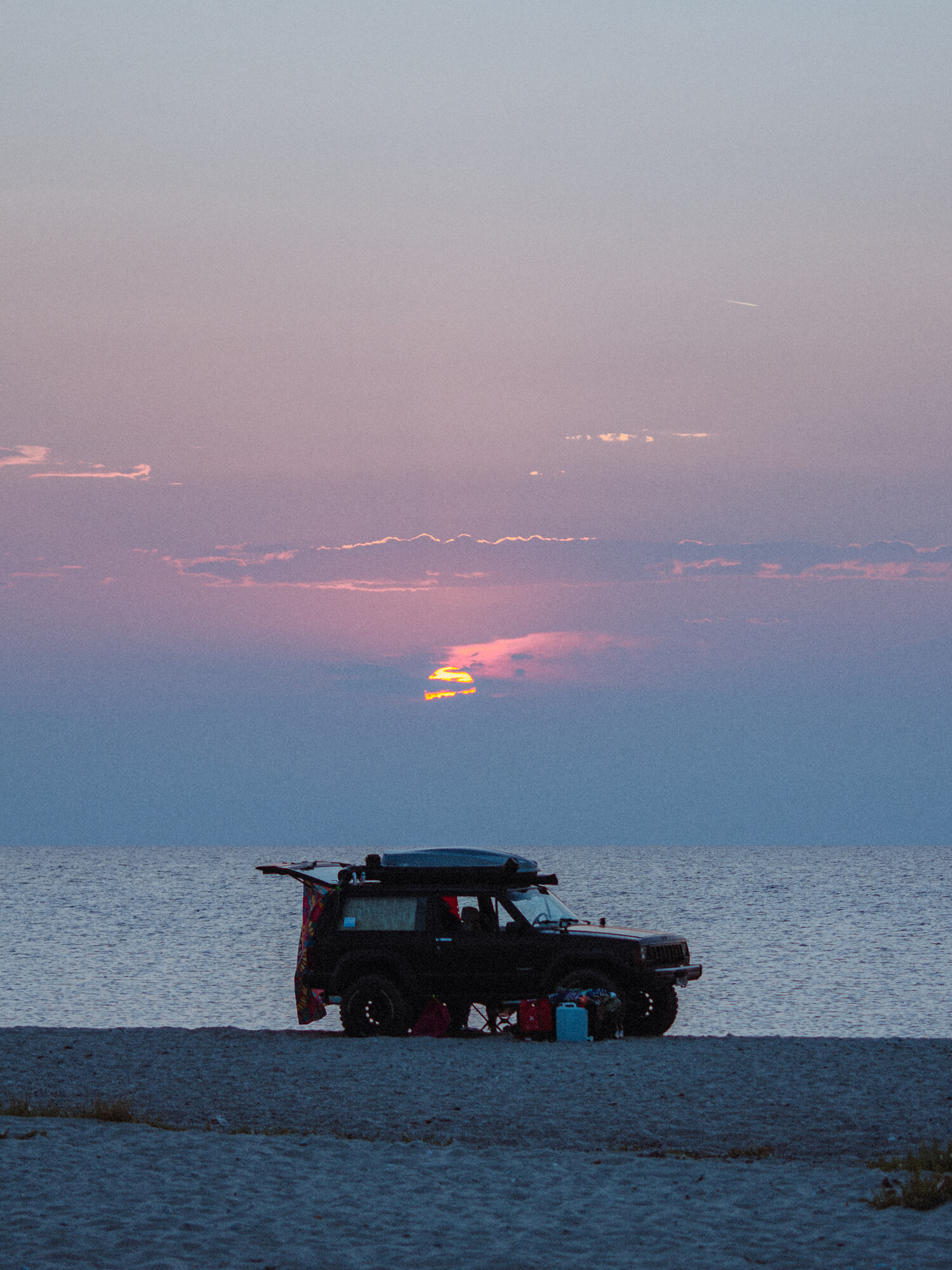 Off-road vehicle parked on a beach silhouetted against a sunset over the sea