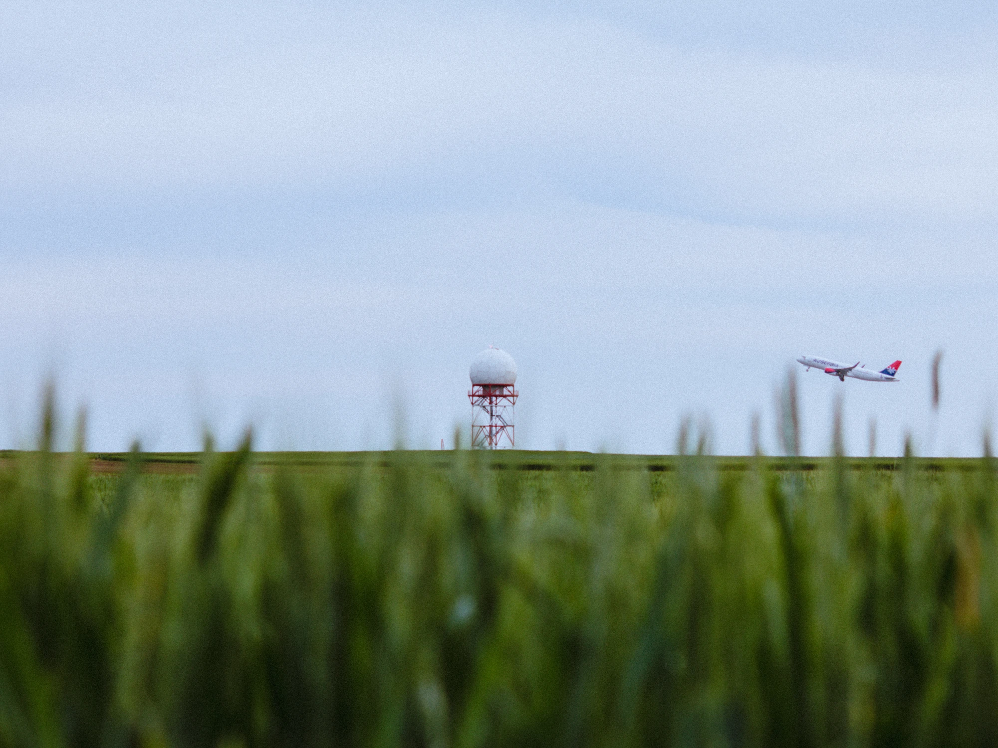 Airplane taking off near radar tower beyond grassy field under cloudy sky