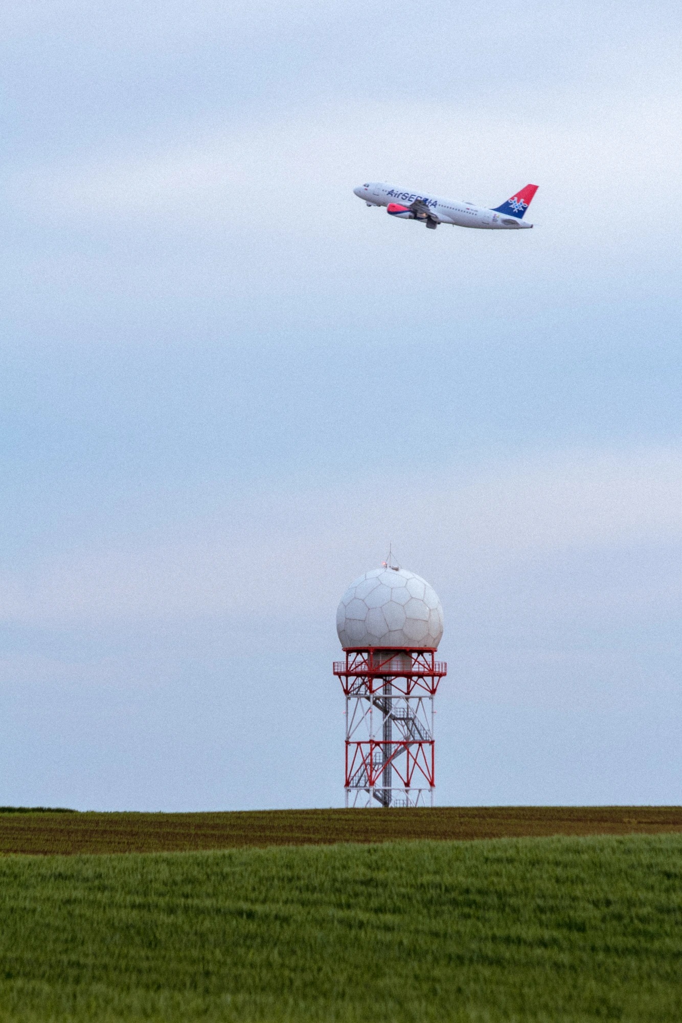 Airplane flying above a radar tower over a grassy field under a cloudy sky