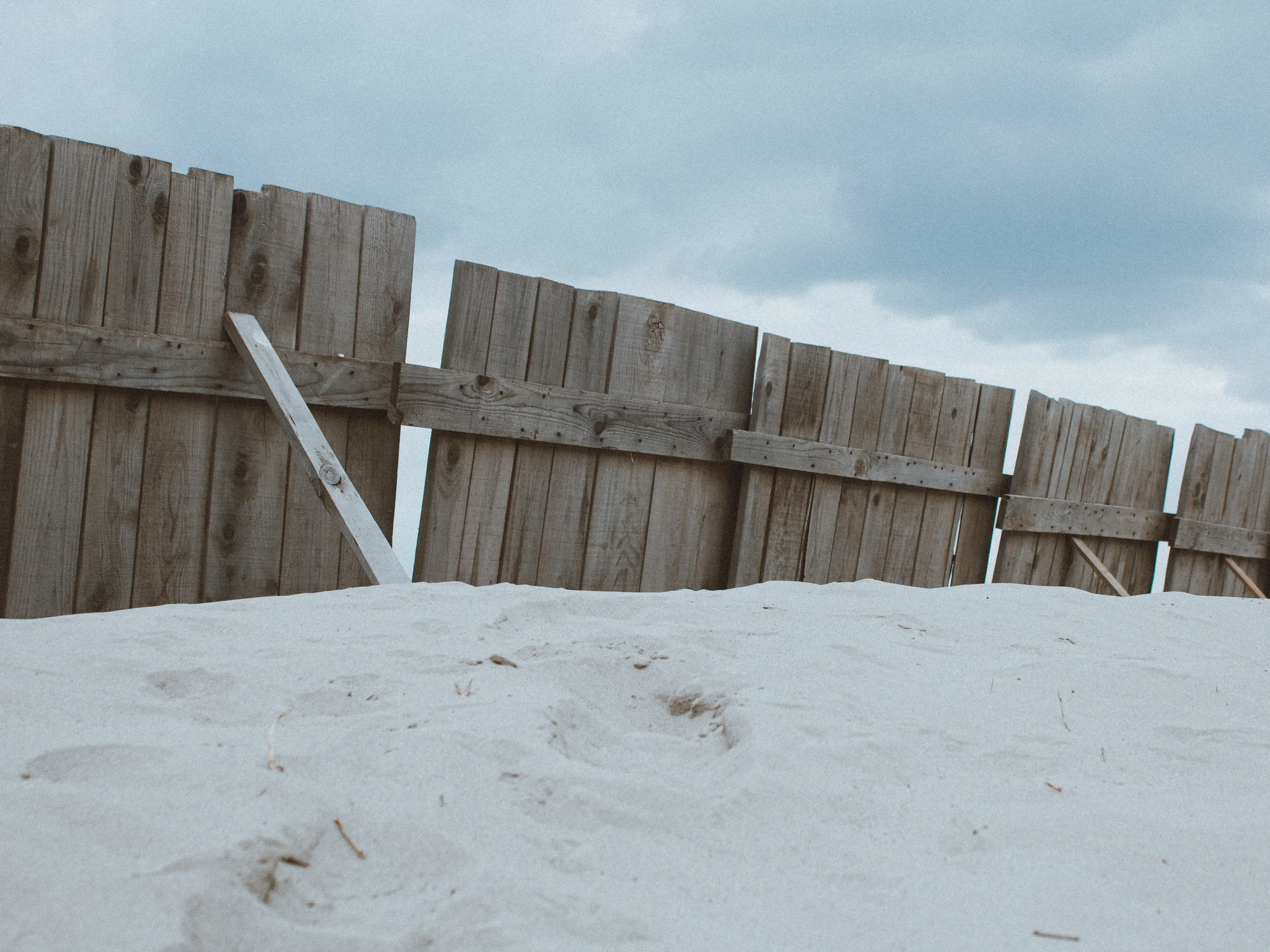 A weathered wooden fence half-buried in sand dunes under a heavy overcast sky