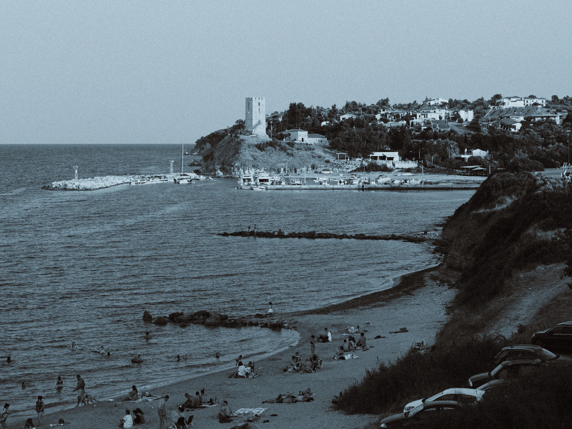 Black and white view of a Greek coastal village with a stone tower overlooking a harbor and beach
