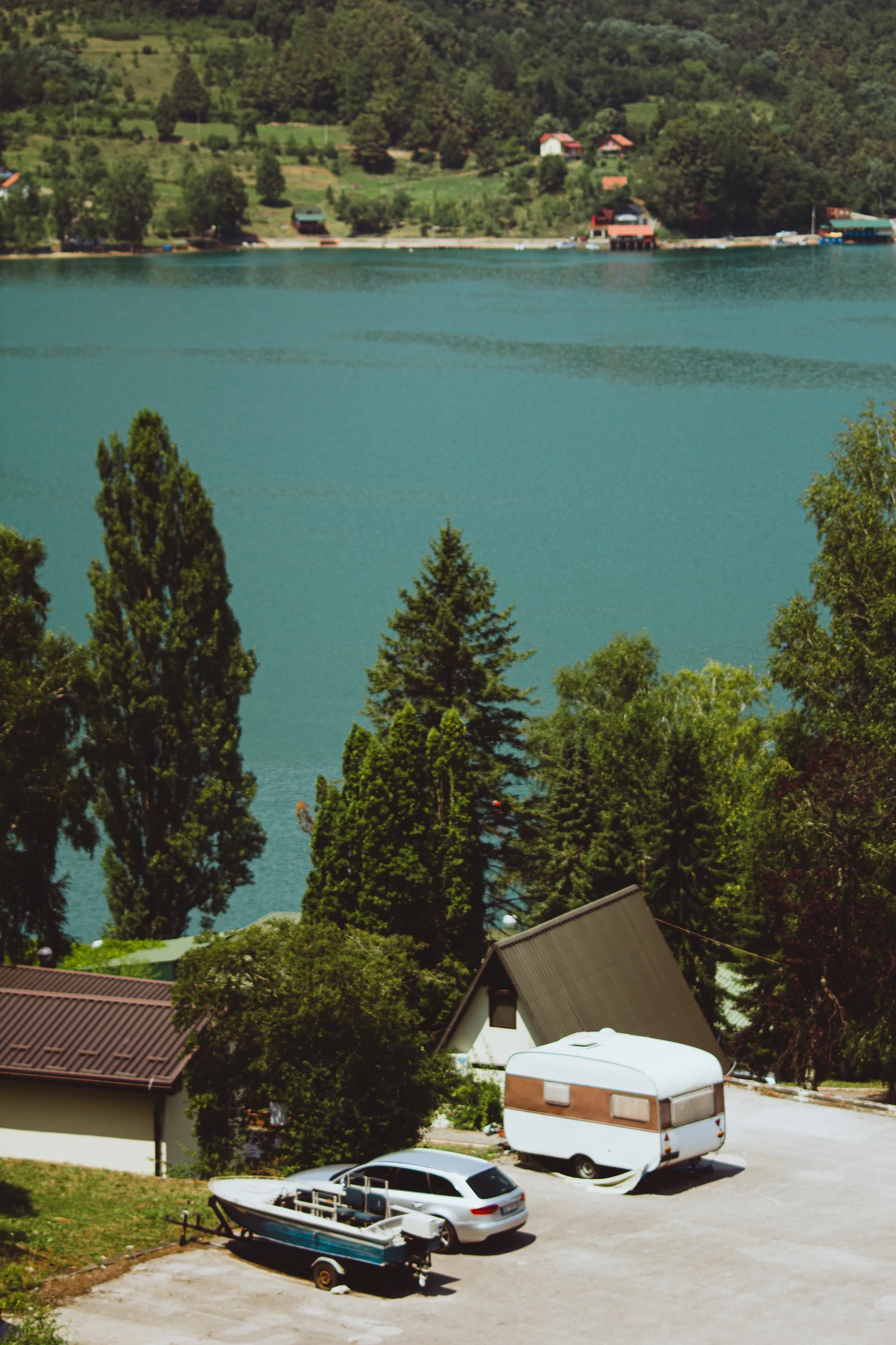 A boat, a car and a travel trailer on a parking with cypress trees and lake in the background